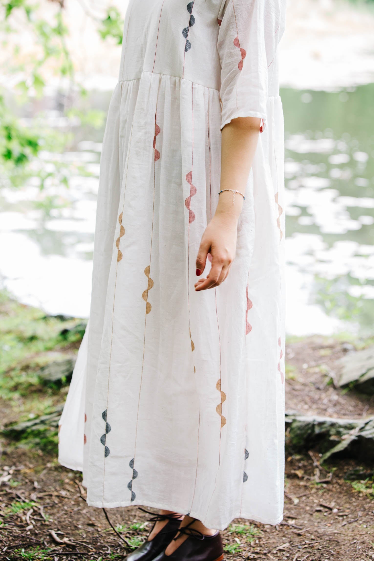 Person wearing a handwoven white jamdani dress with red and black patterns standing near a body of water.