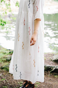 Person wearing a handwoven white jamdani dress with red and black patterns standing near a body of water.
