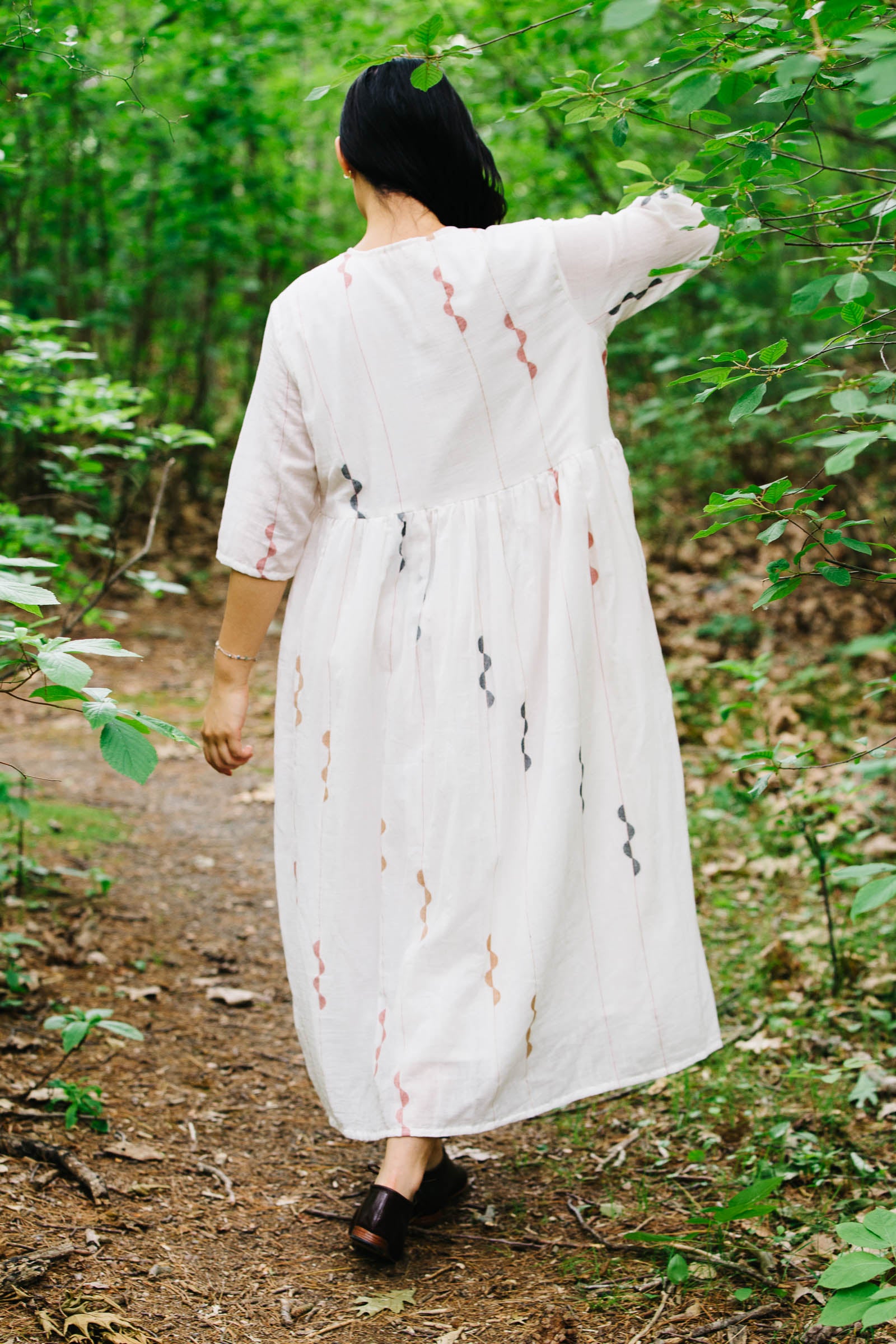 Person wearing an artisan made handwoven white jamdani dress with red and black patterns walking through a forest.