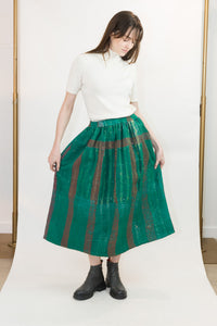 A model wearing a white short-sleeve mock neck top and a voluminous green pleated skirt with vertical red stripes, paired with black ankle boots, standing against a white studio backdrop.