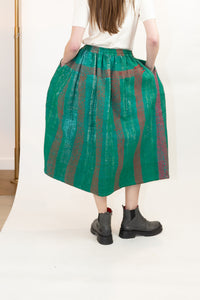 A person shown from the back wearing a full, quilted green skirt with vertical red stripes, paired with a white top and dark gray Chelsea boots, set against a plain white studio background.