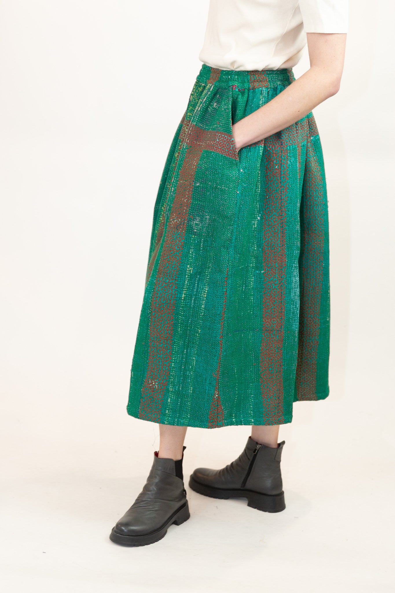 A model is shown from the waist down wearing a high-waisted, wide-leg midi skirt in green with red vertical stripes and a textured, quilted pattern, styled with a white top and black ankle boots against a plain white background.