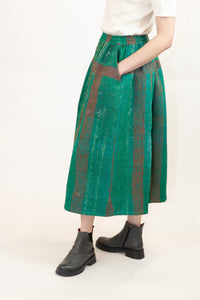A model is shown from the waist down wearing a high-waisted, wide-leg midi skirt in green with red vertical stripes and a textured, quilted pattern, styled with a white top and black ankle boots against a plain white background.