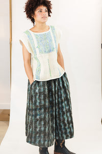A woman modeling a sleeveless knit top with a cream base and light blue and green textured panels, paired with a dark teal and black patterned maxi skirt and black lace-up boots, set against a white studio backdrop.