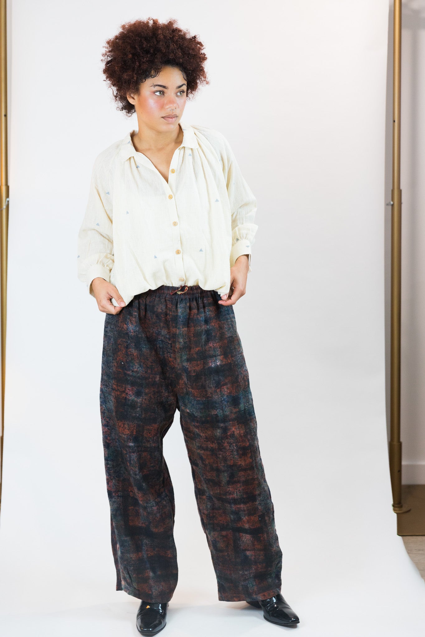A person modeling a cream-colored long-sleeve button-up gather shirt and wide-leg dark plaid trousers with black shoes, standing against a plain white studio background.