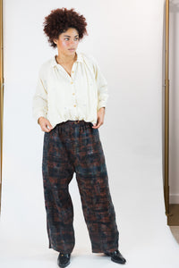 A person modeling a cream-colored long-sleeve button-up gather shirt and wide-leg dark plaid trousers with black shoes, standing against a plain white studio background.