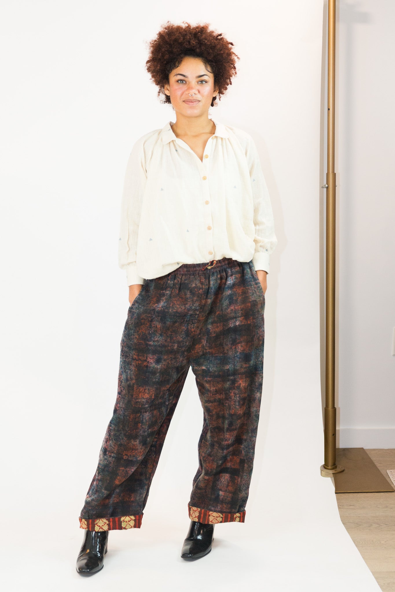 A person modeling loose, wide-leg pants with a dark brown and black abstract pattern, paired with a cream button-up shirt and black boots, standing against a white studio backdrop.