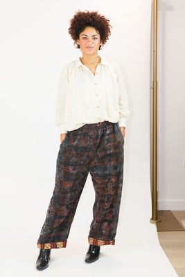 A person modeling loose, wide-leg pants with a dark brown and black abstract pattern, paired with a cream button-up shirt and black boots, standing against a white studio backdrop.