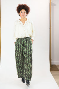 A model stands on a white studio floor wearing a cream-colored long-sleeve button-up gather shirt and green patterned abstract wide-leg trousers, paired with black shoes.