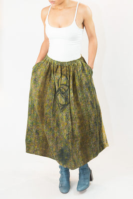 A model wearing a white camisole top and a long, pleated skirt with a green and yellow abstract pattern and side pockets, paired with blue boots, against a plain white background.